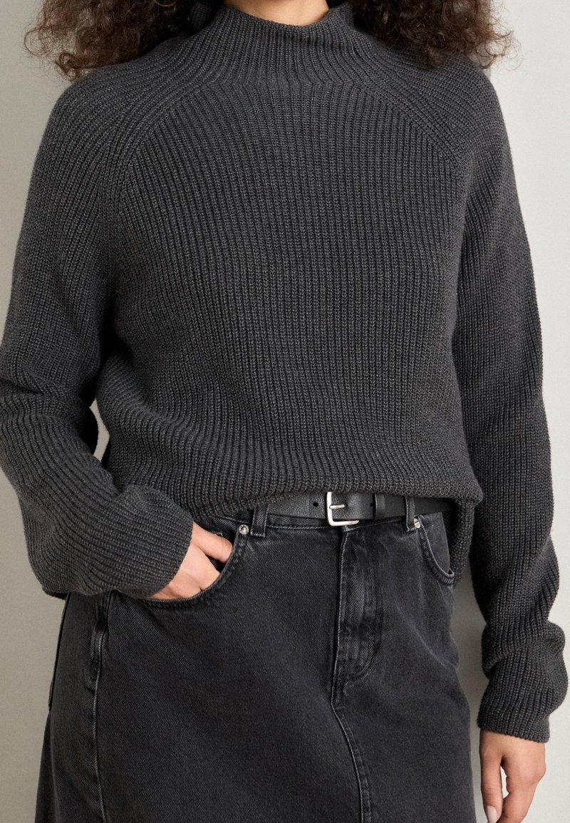 Personne portant un pull en maille côtelée gris foncé rentré dans une jupe en denim noire avec une ceinture noire, main dans la poche de la jupe, sur un fond neutre.