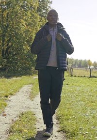 Cilvēks iet pa celiņu, ģērbies tumšā pūkā, pelēkā džemperī un melnās biksēs, ar sporta apaviem; apkārt zaļā zāle un koki.