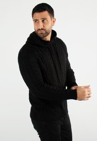 Pull noir en maille torsadée avec capuche, doté de poignets et d'un ourlet côtelés. Conçu pour une coupe ajustée, le motif texturé ajoute un intérêt visuel.