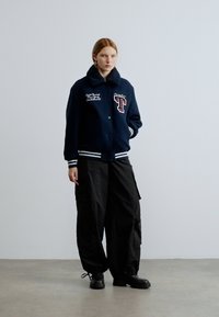 Tamsiai mėlynas universiteto stiliaus džinsinis švarkas su baltomis juostelėmis, ant kurio išsiuvinėta raidė "T". Derinamas su laisvomis juodomis kargo kelnėmis ir stambiais juodais batais.