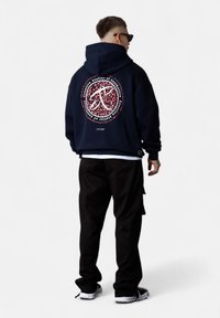 Homme portant un sweat à capuche noir avec un logo circulaire à imprimé léopard rouge et du texte, un pantalon noir, des baskets et des lunettes de soleil, se tenant contre un fond blanc.