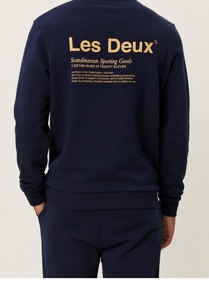 Osoba w granatowej bluzie z beżowym napisem "Les Deux Scandinavian Sporting Goods" oraz opisem na plecach.