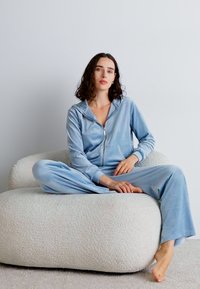 Blå sammetsliknande zip-up jumpsuit med huva, med långa ärmar och vida byxor, sittande på en mjuk, rund krämfärgad stol.