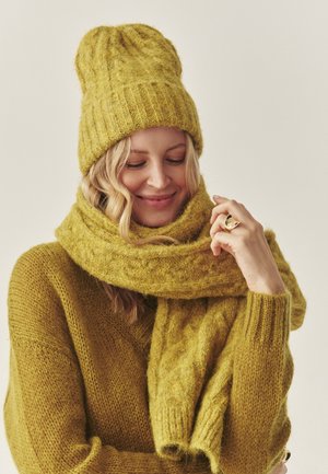 Sárga textúrájú kötött sapka és hozzáillő sál bordázott szélekkel, sárga pulóver felett viselve. Puha, vastag fonal meleg, otthonos megjelenést kölcsönöz.