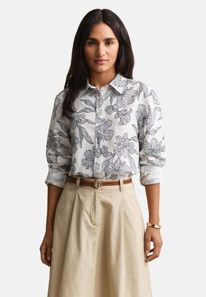 Ženska ima na sebi belo srajco z gumbi, črnim cvetličnim potiskom, bež krilo z visokim pasom, rjav pas in zlato zapestnico, stoji pred belim ozadjem.