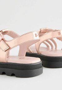 Vaaleanpunaiset metallinväriset sandaalit, joissa on useita hihnoja, sileä varvasalue ja teksturoitu musta kumipohja. Suunnittelussa on pieni logo-ysitys.