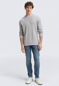 Hellgraues, geripptes Langarmshirt kombiniert mit hellblauen, eng anliegenden Jeans und weißen Turnschuhen, stehend vor einem schlichten Hintergrund.