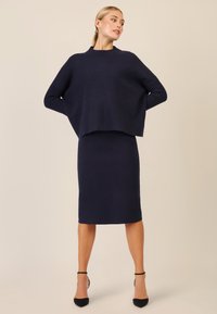 Granatowy sweter dzianinowy o kroju cropped z prążkowanymi rękawami, zestawiony z dopasowaną spódnicą o długości do kolan w kształcie ołówka. Czarne buty na wysokim obcasie.