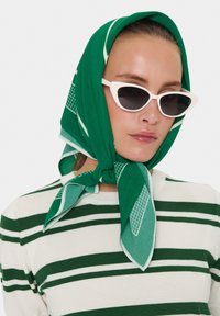 Foulard di seta verde a fantasia annodato intorno al collo, abbinato a un maglione a righe verdi e bianche. La modella indossa occhiali da sole bianchi a forma di occhio di gatto.