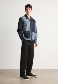 Denimtakki, jossa on päällekkäin ommeltu design vaaleista ja tummista sinisistä kankaista, nappikiinnitys edessä ja kaksi taskua. Yhdistetty mustien housujen ja kenkien kanssa.