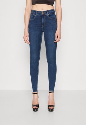 Vrouw draagt donkerblauwe skinny jeans met hoge taille, dubbele houten knopen en zwarte open tenen hoge hakken, staand tegen een witte achtergrond.