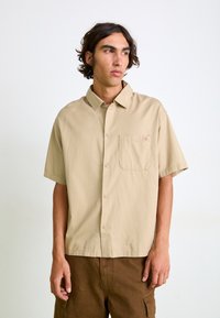 Short-sleeve beige button-up shirt made of lightweight fabric, featuring a chest pocket and collar, with a relaxed fit.