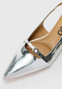 Sidabrinė smailėjanti slingback avalynė su metaliniu paviršiumi, turinti siaurą reguliuojamą dirželį ir lygų, tekstūruotą vidpadį.