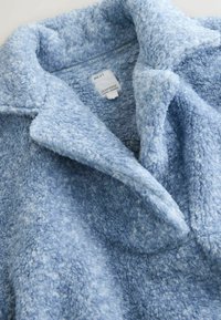 Blå fleecejakke med en blød, struktureret overflade; har en skårede krave og et mærke, der indikerer "downtime" og "NEXT."