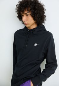 Mlad človek s kodrastimi lasmi, oblečen v črno Nike kapuco z vrvicami, ki stoji ob beli steni, roka v žepu vijoličnih hlač.