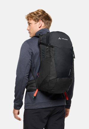 Mann mit dunkelblauer Jacke und schwarzem Wanderrucksack mit Hüftgurt vor weißem Hintergrund.