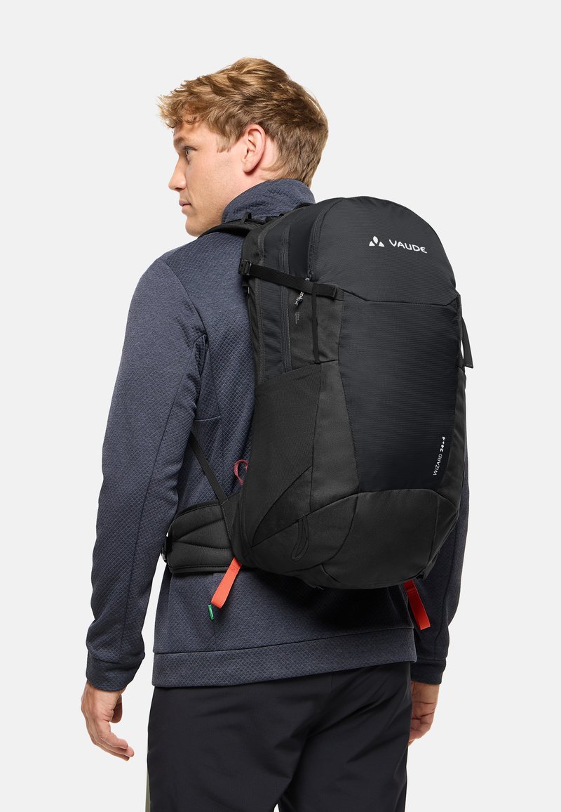 Hombre con chaqueta azul oscuro y mochila de senderismo negra con cinturón de cintura sobre fondo blanco.