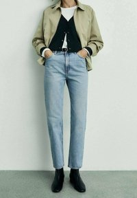 Veste beige légère portée sur un cardigan noir, associée à un jean bleu clair et des bottines noires. Design minimaliste, silhouette décontractée.