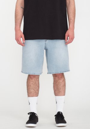 Homme portant un short en jean bleu clair, des chaussettes blanches montantes, des baskets noires et un t-shirt noir, debout sur un fond blanc.