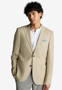 Blazer beige avec un revers cranté, deux poches avant et deux boutons. Porté par-dessus une chemise de couleur claire avec un mouchoir de poche à motifs.