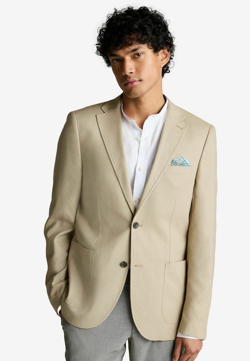 Blazer beige avec un revers cranté, deux poches avant et deux boutons. Porté par-dessus une chemise de couleur claire avec un mouchoir de poche à motifs.