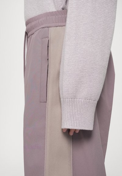 Calças de treino lilás claras com cintura elástica, painel lateral bege em contraste e bolso com fecho zip. Textura suave e ajuste confortável.