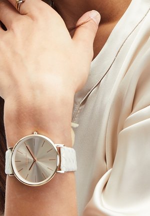 Poignet portant une montre argentée avec un bracelet matelassé blanc, main touchant le cou, portant un chemisier en satin crème.