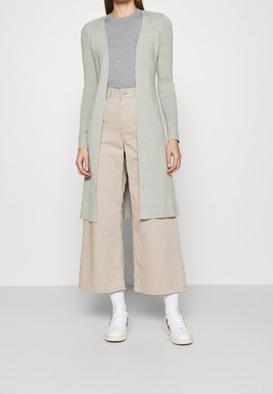 Femme portant un long cardigan ouvert gris clair, un t-shirt gris, un pantalon large beige, des chaussettes blanches et des baskets blanches, debout devant un fond uni.