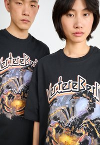Black cotton T-shirts feature a bold graphic print of a scorpion with colorful details. The design includes stylized text above the image.