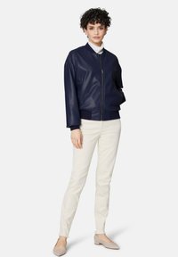 Marine faux leren bomberjack met ribgebreide mouwen en onderrand, ritssluiting, gedragen met lichtgekleurde broek en platte schoenen. Eenvoudig, strak ontwerp.