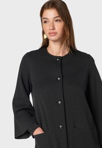 Giacca nera con bottoni, colletto rotondo, maniche lunghe e due tasche frontali. Realizzata in un tessuto strutturato, presenta grandi bottoni neri.