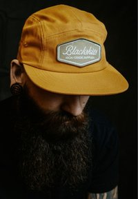 Kollane puuvillane müts lapikuga äärega ja ristkülikukujuline silt, millel on valged kirjad "Blackskies" ja "HIGH-GRADE SUPPLIES".