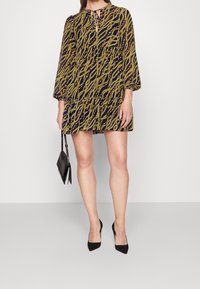 Lyhythihainen mini mekko, jossa on tummansininen pohja ja keltainen aaltoileva kuvio. Kaula-aukossa on avainaukko-yksityiskohta. Yhdistetty mustiin kärkitossuihin ja pieneen käsilaukkuun.