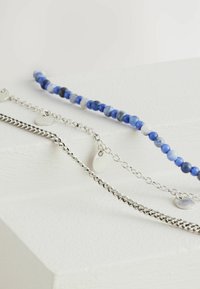 Tre armbånd: et armbånd med blå perler, en tynd sølvkæde med cirkulære accenter og en struktureret snoet sølvkæde.