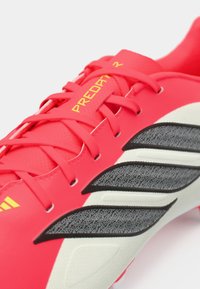 Κόκκινα ποδοσφαιρικά παπούτσια με μαύρες και λευκές υφασμάτινες λεπτομέρειες, που διαθέτουν απαλό επάνω μέρος, κλείσιμο με κορδόνια και κίτρινο κείμενο "Predator R".