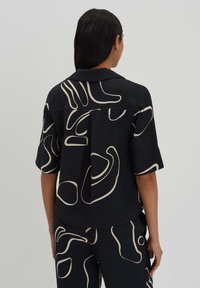 Chemise noire à manches courtes texturée avec un motif abstrait beige. Le dos comporte un pli central pour plus de mouvement et de détail.