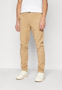 Chino khaki realizzati in cotone, con una vestibilità slim, tasche anteriori visibili e chiusura con un singolo bottone, abbinati a sneakers bianche.
