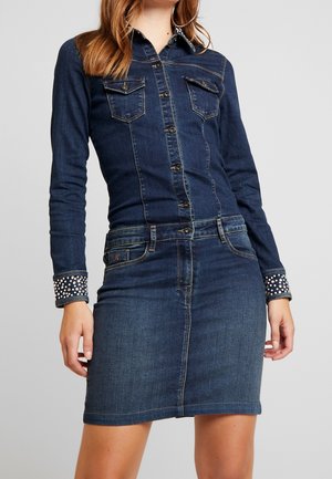 Femme portant une veste en jean bleu foncé avec des poignets cloutés et une jupe en jean assortie sur fond uni.