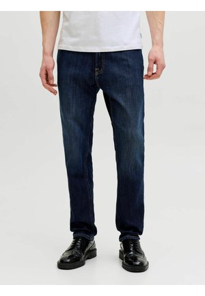 Mann trägt dunkelblaue, schmal geschnittene Jeans, weißes Hemd und schwarze Leder-Schnürschuhe, steht vor hellem Hintergrund.