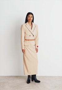 Beige kurzes Blazerjackett mit schwarzen Akzenten, kombiniert mit einem passenden langen Rock. Das Modell trägt schwarze Stiefel mit Absätzen. Glatte Textur, formelles Design.