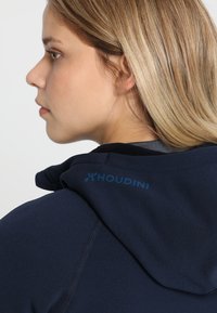 Navy sininen huppari, jossa on pehmeä pinta ja selässä kaulan lähellä näkyvä sininen "Houdini"-logo. Yksinkertainen ja toiminnallinen muotoilu.