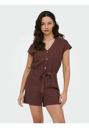 Kvinne med mørkt, bølgete hår iført brun kortermet romper med knapper og belte i midjen, stående mot en ensfarget lys bakgrunn.