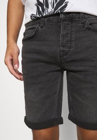 Hombre vestido con pantalones cortos de mezclilla negros con dobladillo enrollado y una camiseta blanca con impresión gráfica negra, de pie con la mano izquierda relajada a un lado.