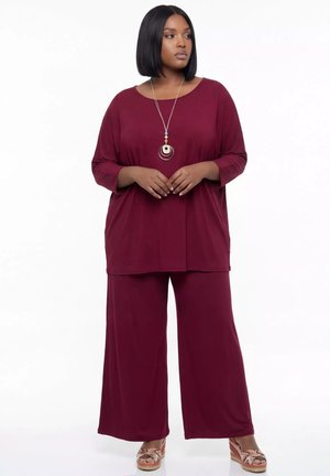 Kobieta ubrana w luźny bordowy top i spodnie z szerokimi nogawkami, z długim naszyjnikiem i sandałami na koturnie, stojąca na białym tle.