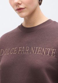 Barna pulóver bordázott kerek nyakkivágással, a mellkason bézs hímzett "DOLCE FAR NIENTE" felirattal. Simább textúrájú.