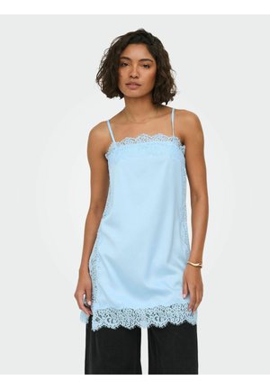 Femme portant une tunique sans manches bleu clair bordée de dentelle sur un pantalon noir à jambes larges, debout devant un fond clair uni.