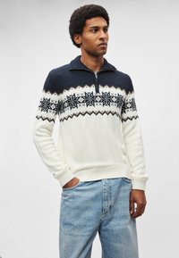 Sweter dziany w kremowym kolorze z granatowym prążkowanym kołnierzem, ozdobiony geometrycznym wzorem płatka śniegu na klatce piersiowej oraz kontrastującymi mankietami.