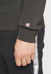Primo piano di una mano accanto a un polsino di felpa scura con un logo Champion rosso e bianco, e pantaloni neri con una striscia del logo Champion bianca.
