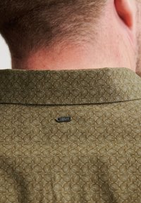 Camicia verde oliva con un motivo geometrico testurizzato. Colletto a vista con una piccola etichetta metallica del marchio sul retro.
