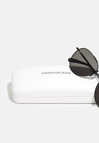Calvin Klein Jeans solbriller med en slank svart ramme og mørke linser, som hviler på et hvitt rektangulært etui med en glatt overflate og logo.